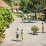 Unser Hof lädt zum Verweilen ein, © Winzerhof Bogner Ein sonniger Hof mit Menschen, die spazieren, Tischtennis spielen und sich unterhalten. Umgeben von Pflanzen und Gebäuden mit roten Dächern.