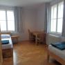 Gästezimmer, © Geistliches Jugendzentrum Oberleis Ein einfach eingerichtetes Gästezimmer mit zwei Betten, einem Tisch und Stühlen, Holzboden und großen Fenstern mit Vorhängen.