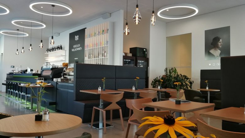 Poldi Fitzka Innenbereich, © Roman Zöchlinger Innenansicht eines modernen Cafés mit runden Deckenleuchten, Holztischen und Blumen auf den Tischen.