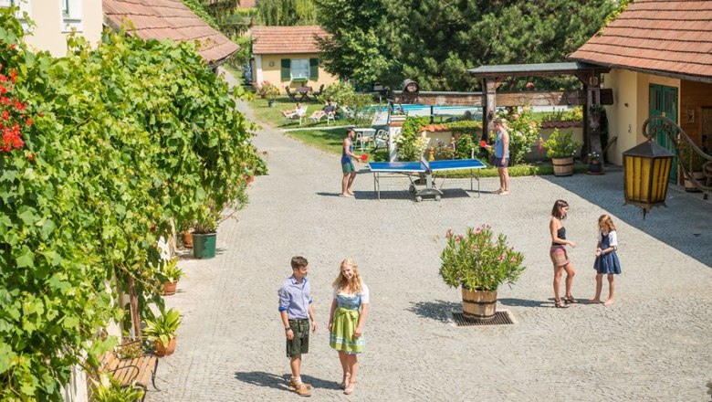 Our farm invites you to linger, © Winzerhof Bogner A sunny courtyard with people walking, playing table tennis and chatting. Surrounded by plants and buildings with red roofs.