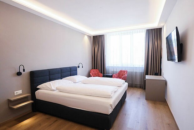 Sample room, © Parkhotel Modern hotel room with a double bed, two red armchairs, a large window and a flat-screen TV on the wall.