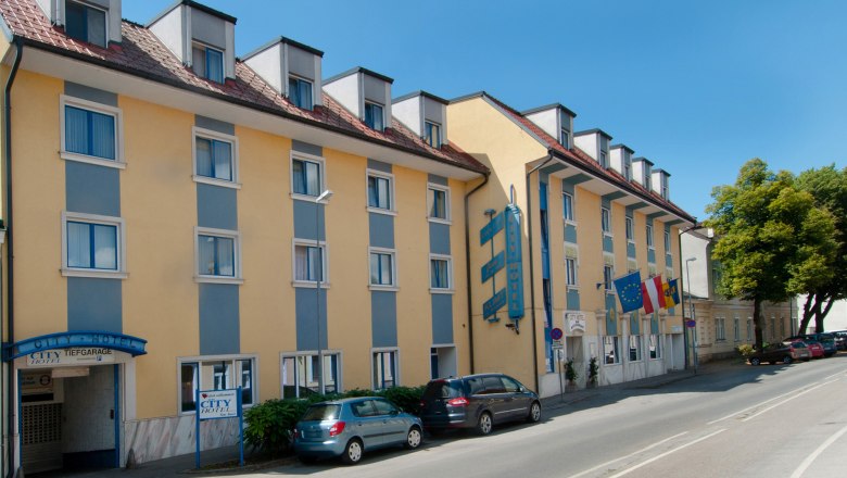 Pohled na exteriér hotelu City, © © Markus Jung, MJM Photography Pohled zvenčí na žlutomodrý hotel s několika okny a zaparkovanými auty před ním.