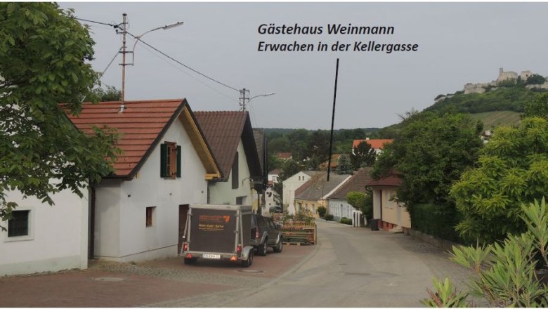 Kellergasse, © Fam. Weinmann Eine malerische Straße mit traditionellen Häusern und einem Anhänger. Im Hintergrund ist eine Burg auf einem Hügel zu sehen.