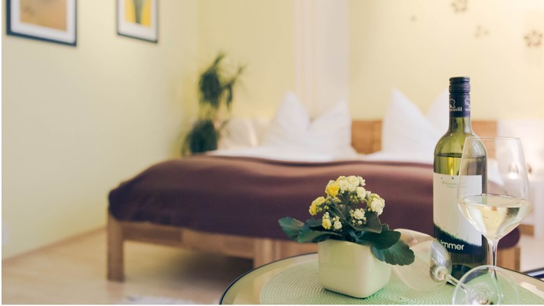 A cozy bedroom with a bed in the background and a table with a bottle of wine and a glass in the foreground.