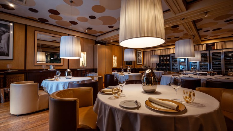 Elegantn&iacute; restaurace s kulat&yacute;mi stoly, b&iacute;l&yacute;mi ubrusy a velk&yacute;mi z&aacute;věsn&yacute;mi lampami.