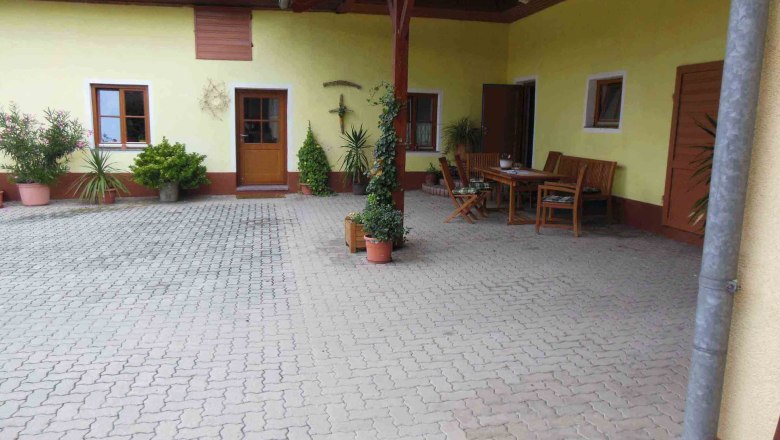 Innenhof mit gelber Fassade, Holztisch und Stühlen, umgeben von Pflanzen in Töpfen.