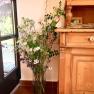 Ein Strauß wilder Blumen in einer Vase neben einem Holzmöbelstück, mit Blick auf einen Garten durch ein Fenster.