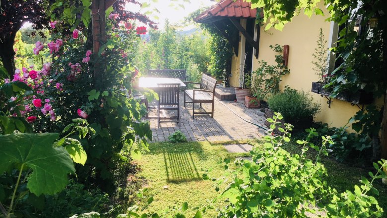Ein idyllischer Garten mit blühenden Rosen, einem Tisch und Stühlen auf einer Terrasse neben einem gelben Haus.
