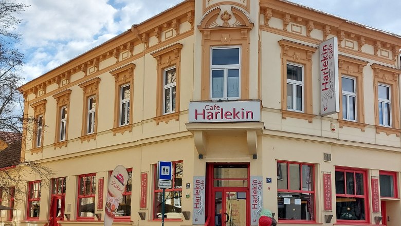 Pohled zvenč&iacute; na kav&aacute;rnu Restaurant Harlekin se žlutou fas&aacute;dou a červen&yacute;mi okny.