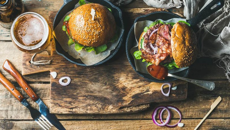 Two burgers in pans with beer and cutlery on a wooden table.