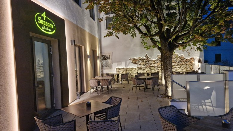 Venkovn&iacute; terasa restaurace se stoly a židlemi, osvětlen&aacute; za soumraku, v pozad&iacute; strom.