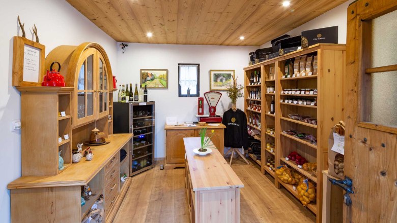 Innenansicht eines kleinen, gemütlichen Ladens mit Holzregalen, gefüllt mit regionalen Produkten wie Wein, Marmelade und Gemüse.