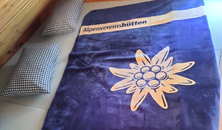Ein Matratzenlager mit einer blauen Decke, die ein Edelweiß und den Schriftzug 'Alpenvereinshütten' zeigt, sowie zwei karierte Kissen.