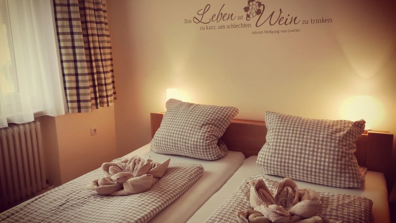 Gem&uuml;tliches G&auml;stezimmer mit zwei Betten, karierten Bettdecken und Kissen, dekorativen Handt&uuml;chern und einem Goethe-Zitat an der Wand.