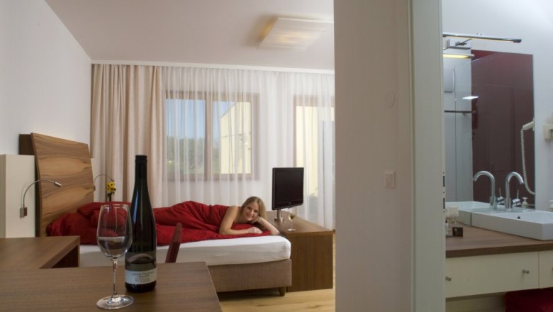 Ein modernes Hotelzimmer mit einem Bett, auf dem eine Person liegt, und einem Tisch mit einer Weinflasche und einem Glas.