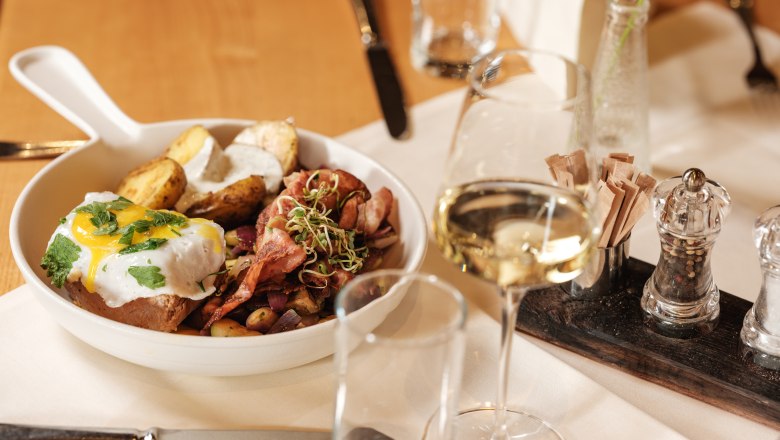 Vřelé přivítání, © Weinviertel Tourismus GmbH / Michael Reidinger Talíř s pokrmem z masa, brambor a smaženého vejce, podávaný se sklenkou bílého vína na prostřeném stole.