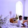 A boy sits on a bed in a bright room with floral curtains and bed linen.