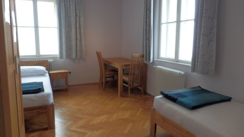 Ein einfach eingerichtetes Gästezimmer mit zwei Betten, einem Tisch und Stühlen, Holzboden und großen Fenstern mit Vorhängen.