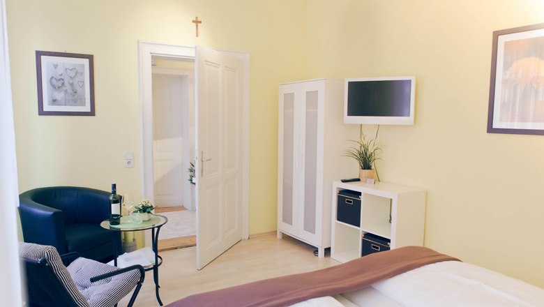 Bright guest room with bed, armchair, table, wardrobe and TV.
