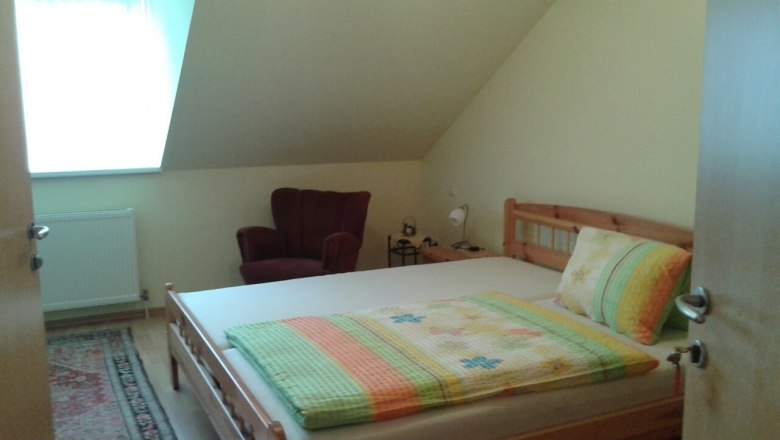 Ein gemütliches Schlafzimmer mit einem Doppelbett, einem roten Sessel und einem kleinen Nachttisch unter einer Dachschräge.