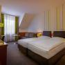 Modernes Hotelzimmer mit zwei Einzelbetten, grünen Lampen und einem Schreibtisch. Fenster mit Vorhängen im Hintergrund.