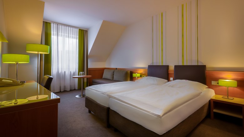 Modernes Hotelzimmer mit zwei Einzelbetten, grünen Lampen und einem Schreibtisch. Fenster mit Vorhängen im Hintergrund.