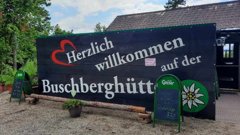 Willkommensschild der Buschberghütte mit grüner Umgebung und blauen Himmel.