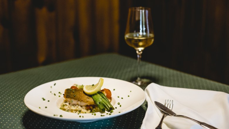 Ein Teller mit Lachsforelle und Risotto, garniert mit Zitrone und Gemüse, steht auf einem gedeckten Tisch neben einem Glas Weißwein.