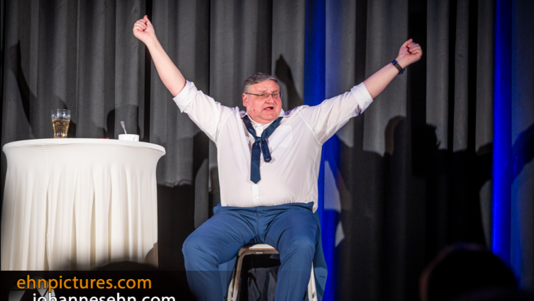 Na p&oacute;diu sed&iacute; muž se zdvižen&yacute;ma rukama, vedle něj je stůl se sklenic&iacute;.