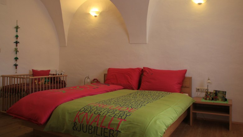 Ein gem&uuml;tliches G&auml;stezimmer mit einem Doppelbett, bunten Bettbez&uuml;gen und einem Babybett im Hintergrund. Die W&auml;nde sind wei&szlig; und gew&ouml;lbt.