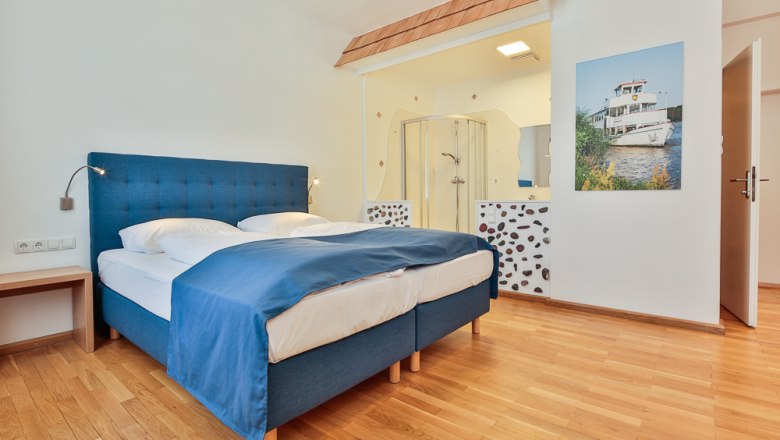 Hotelzimmer mit blauem Bett, Holzboden und offener Dusche.