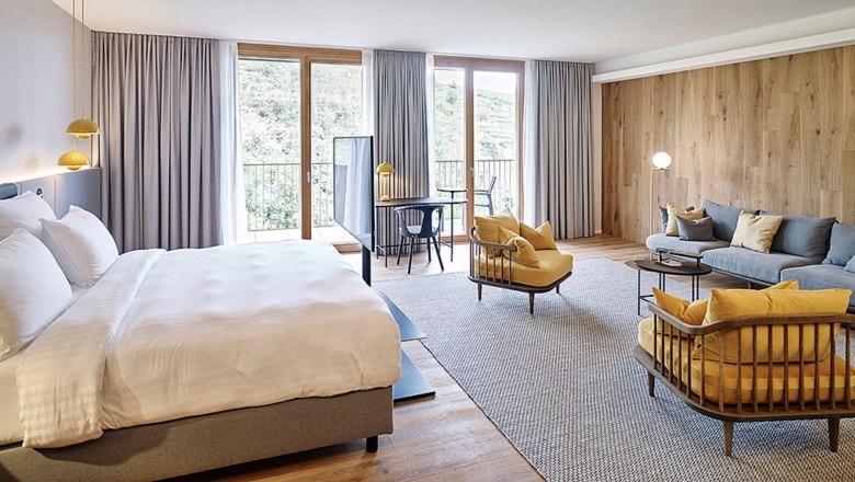 Modernes Hotelzimmer mit großem Bett, Sitzbereich und Schreibtisch vor großen Fenstern.