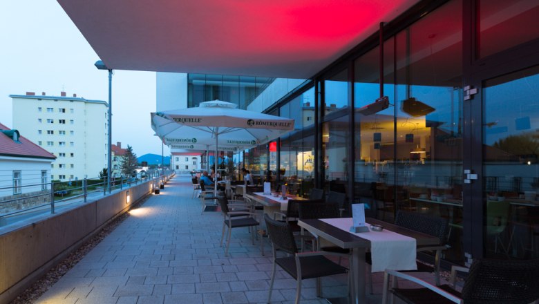 Terrasse eines Restaurants mit Tischen und Stühlen, beleuchtet von roten Lichtern, mit Blick auf Gebäude im Hintergrund.