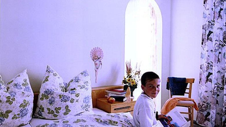 Ein Junge sitzt auf einem Bett in einem Gästezimmer mit Blumenbettwäsche und hält ein Buch. Neben ihm steht ein Nachttisch mit Blumen und Büchern.