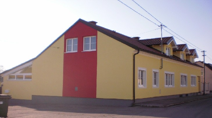 Außenansicht eines gelb-roten Gebäudes mit mehreren Fenstern und einem Satteldach.