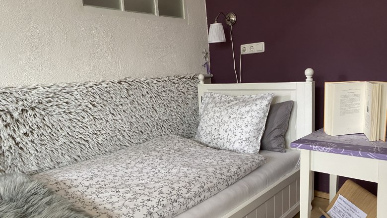 &Uacute;tuln&aacute; ložnice s jednolůžkem, fialovou stěnou, nočn&iacute;m stolkem s knihou a lampou.