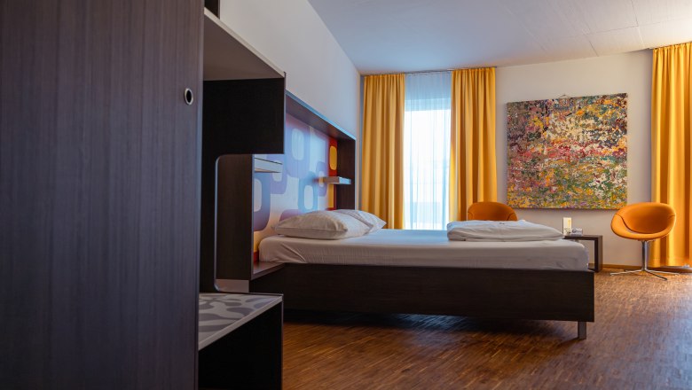 Modernes Hotelzimmer mit Bett, gelben Vorhängen und buntem Gemälde an der Wand.