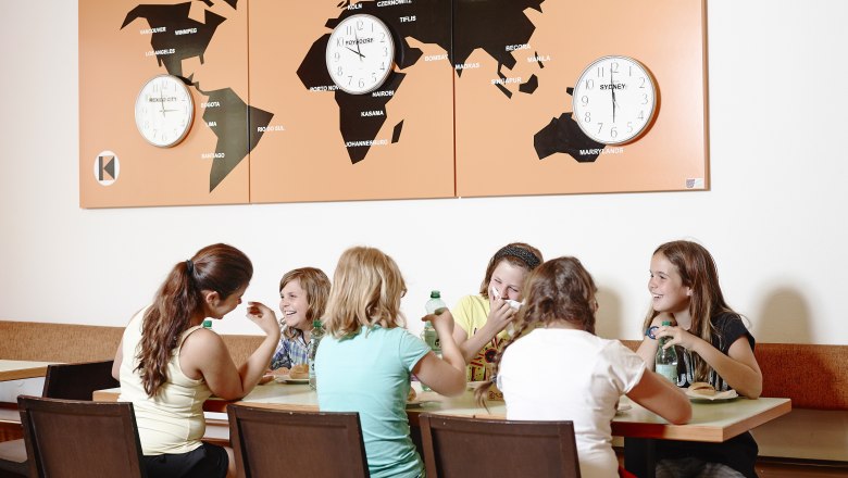 Young people sit at a table and eat, world maps with clocks in the background.
