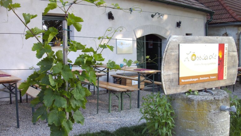 Gastgarten der L&ouml;ssiade mit Holzb&auml;nken, Weinreben im Vordergrund und Fass-Schild vor einem traditionellen Geb&auml;ude bei Tageslicht.