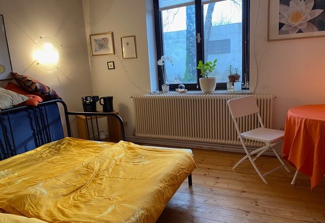 &Uacute;tuln&aacute; ložnice se žlut&yacute;m ložn&iacute;m pr&aacute;dlem, stolem s oranžov&yacute;m ubrusem, rostlinami na parapetu a n&aacute;stěnn&yacute;mi dekoracemi.