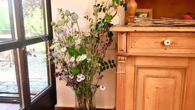 Ein Strauß wilder Blumen in einer Vase neben einem Holzmöbelstück, mit Blick auf einen Garten durch ein Fenster.