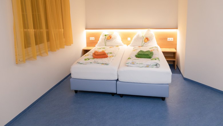 Ein Doppelzimmer mit zwei Einzelbetten, gelben Vorhängen und blauer Boden. Die Betten sind mit Blumenbettwäsche und farbigen Handtüchern dekoriert.
