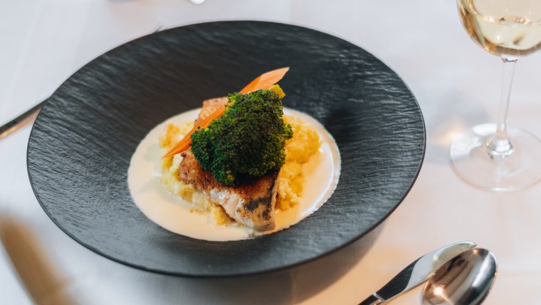 An elegant dish on a black plate with vegetables and sauce, served on a laid table with a glass of white wine.