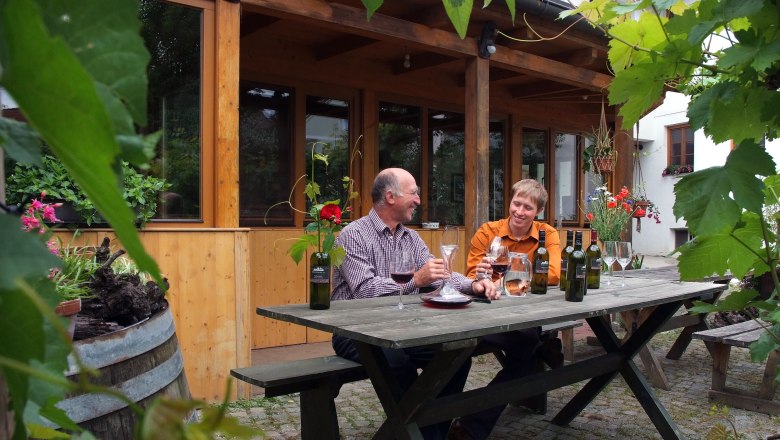 Zwei Männer sitzen an einem Holztisch im Freien und verkosten Wein. Umgeben von Pflanzen und einem Holzgebäude im Hintergrund.