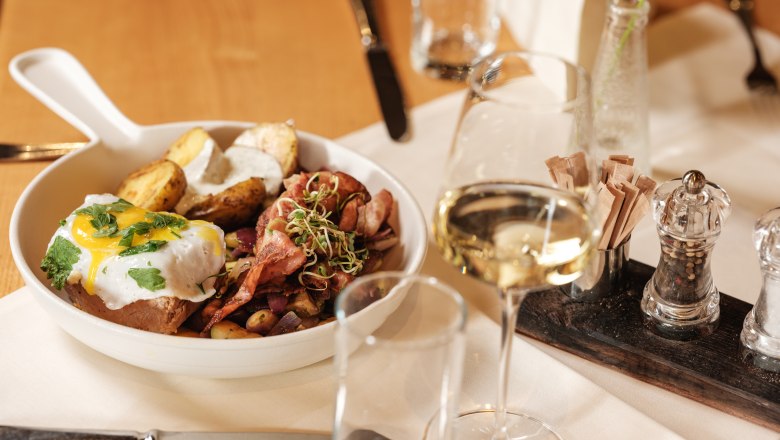 Ein Teller mit Fleisch, Spiegelei, Kartoffeln und Sprossen, daneben ein Glas Weißwein auf einem gedeckten Tisch.