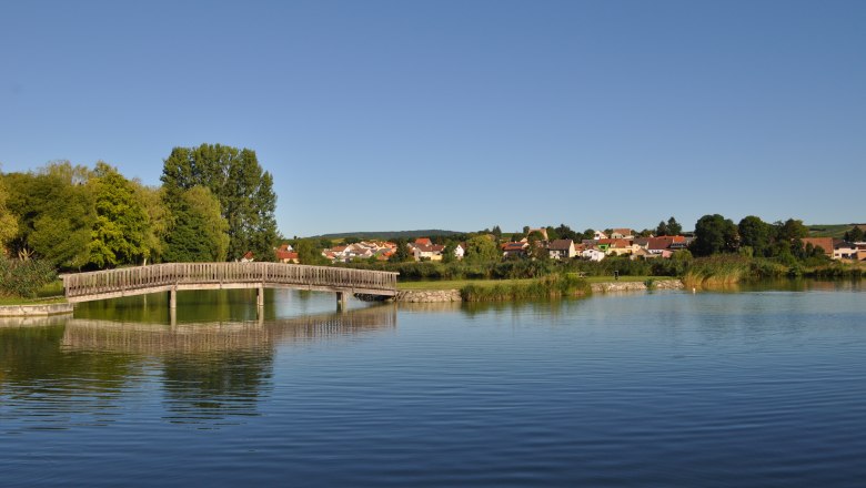 Holzbr&uuml;cke &uuml;ber einen Teich mit Dorf im Hintergrund.