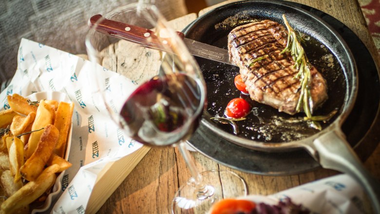 Ein Steak in einer Pfanne mit Tomaten und Rosmarin, daneben Pommes und ein Glas Rotwein auf einem Holztisch.
