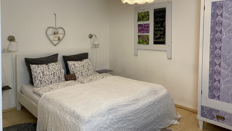 &Uacute;tuln&yacute; dvoulůžkov&yacute; pokoj s b&iacute;lou postel&iacute;, ozdobnou polic&iacute; se srdcem a fialovou &scaron;atn&iacute; skř&iacute;n&iacute;.