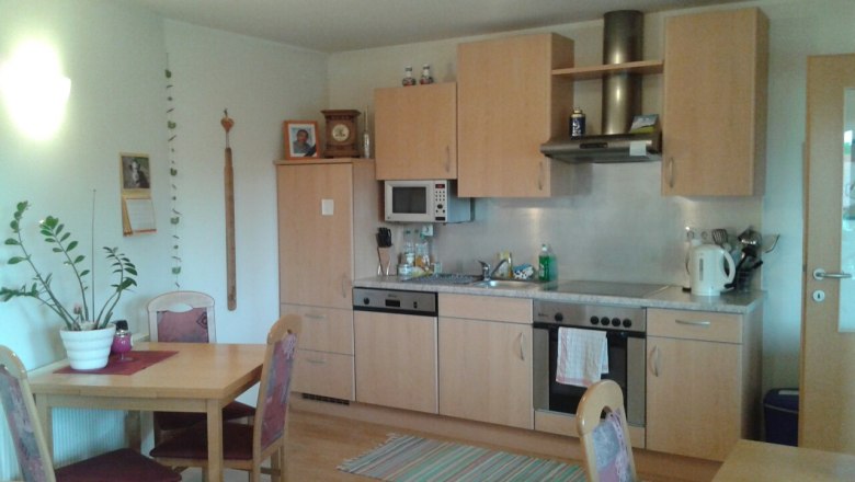 Eine gemütliche Küche mit Holzmöbeln, Esstisch, Stühlen, Spüle, Herd und Mikrowelle. An der Wand hängen Bilder und ein Kalender.