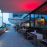 Terrasse eines Restaurants mit Tischen und Stühlen, beleuchtet von roten Lichtern, mit Blick auf Gebäude im Hintergrund.